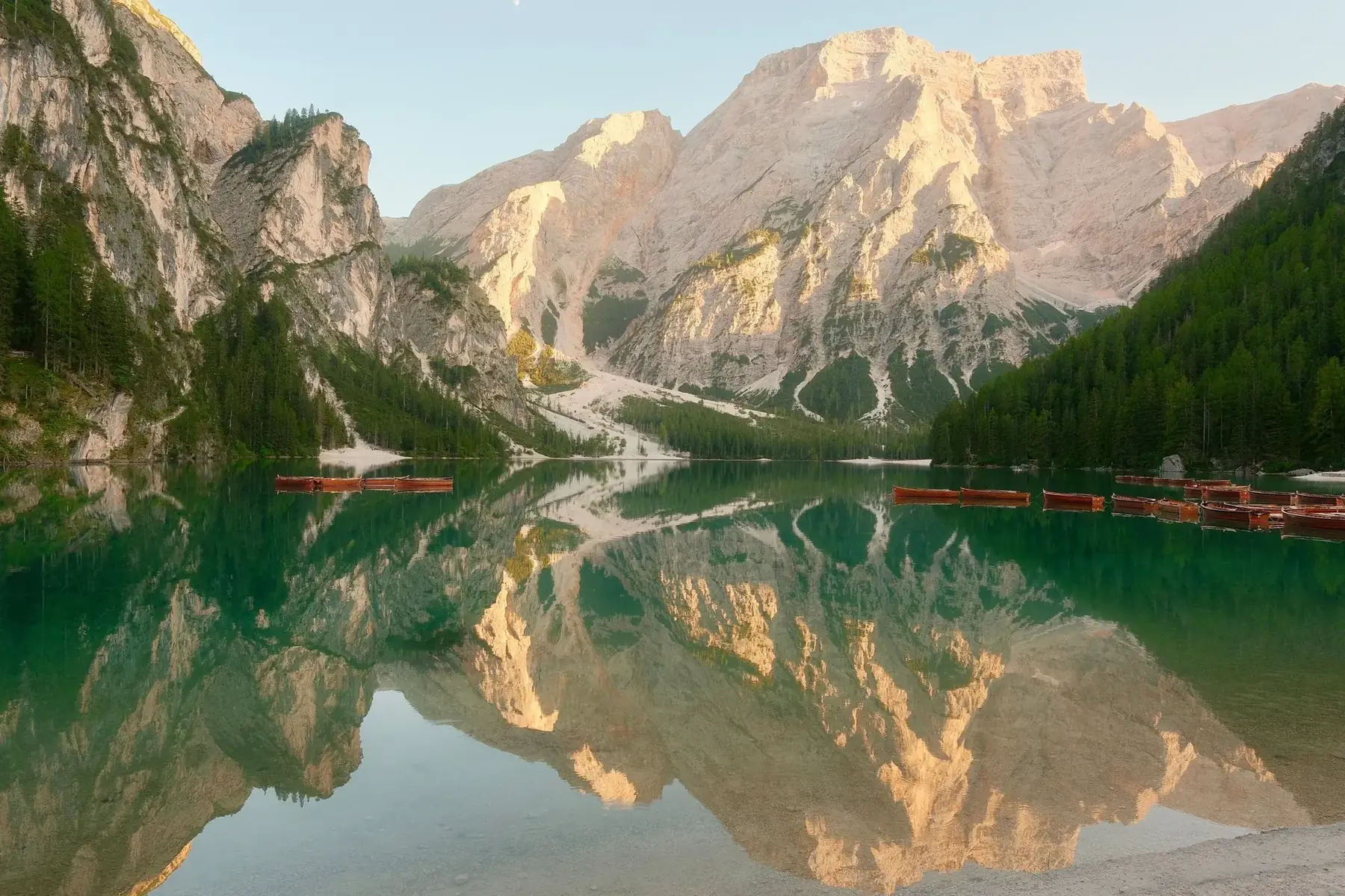 Lago di Braies in warm low light, pale limestone peaks and dark pine slopes mirrored in still green water, a row of red wooden rowboats moored along the shore, Italian Dolomites