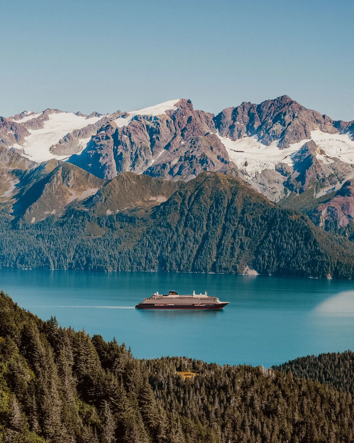 A glacier front in an Alaskan fjord, ice calving into still grey water below forested slopes