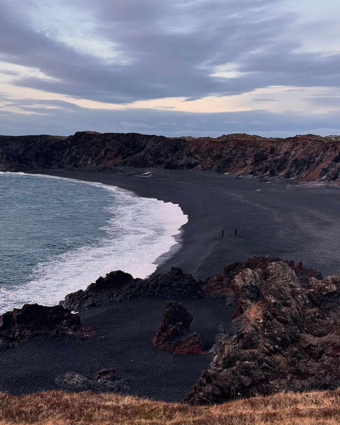 A curving black sand beach in Iceland seen from above, white surf breaking along the bay with dark volcanic rock and a headland beyond