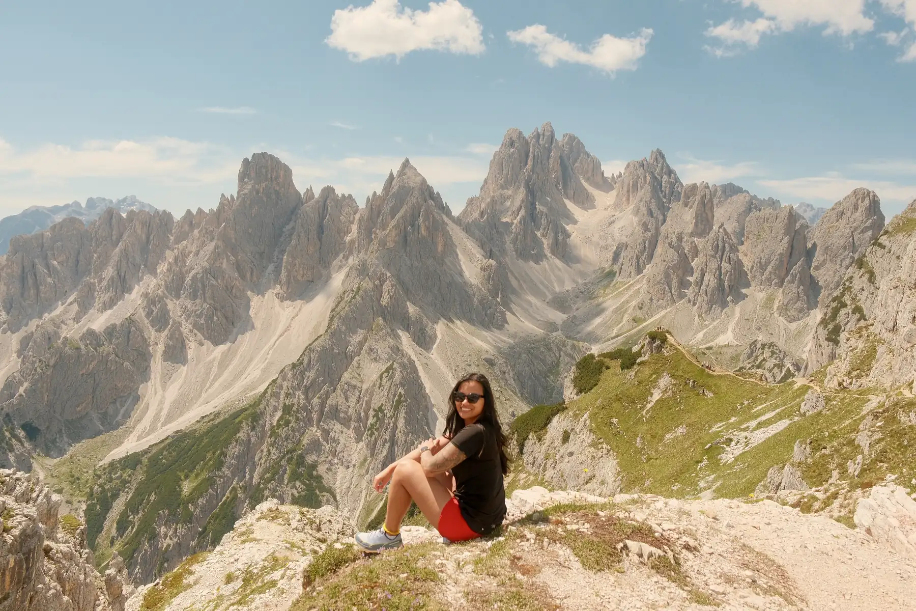 Jea Breshears sitting on a rocky ridge in sunglasses with her arms around her knees, the jagged limestone spires of the Cadini di Misurina filling the sky behind her, Italian Dolomites